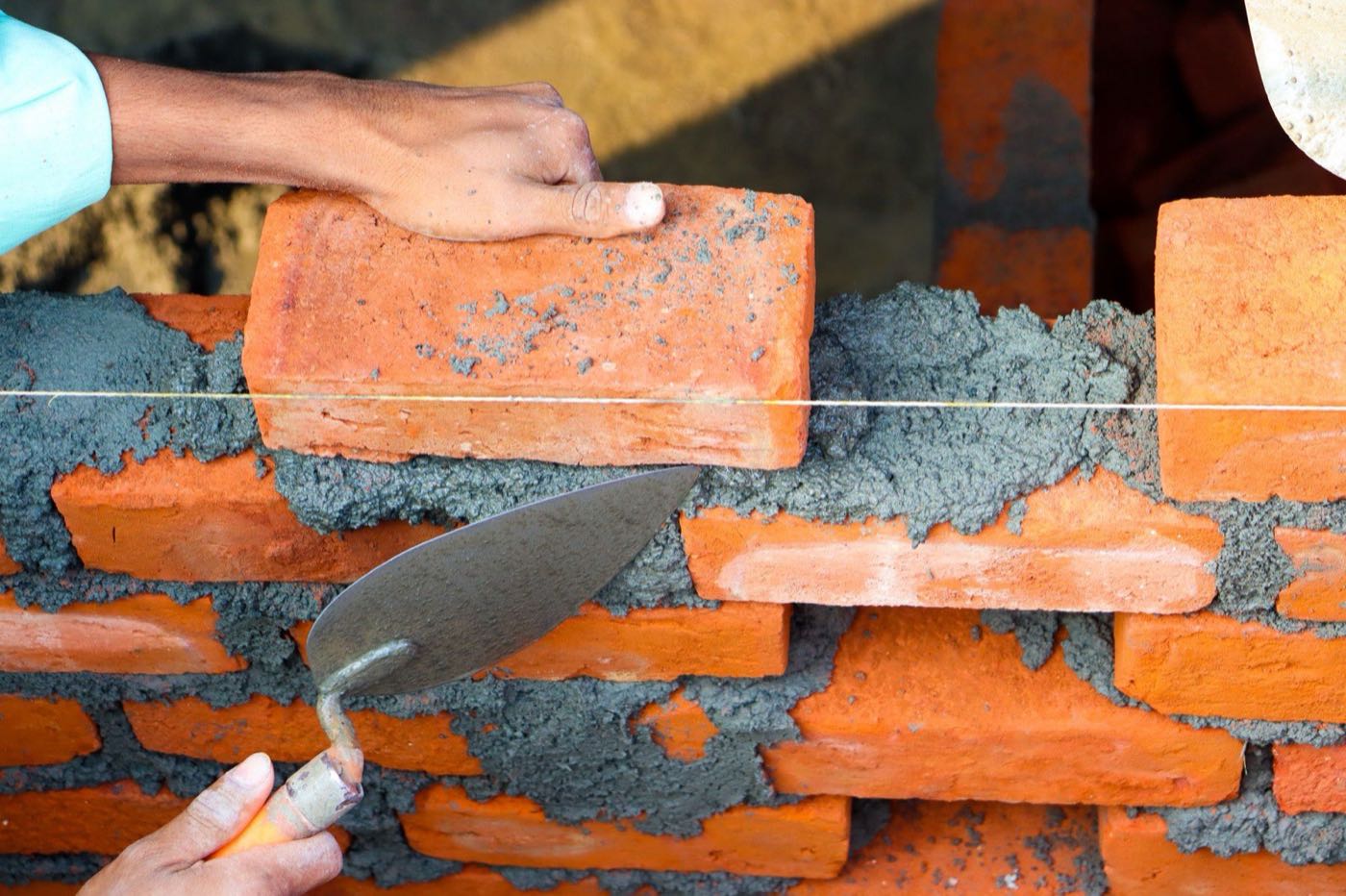 A mason bedding a red clay brick into fresh mortar with a trowel, working to a string line.
